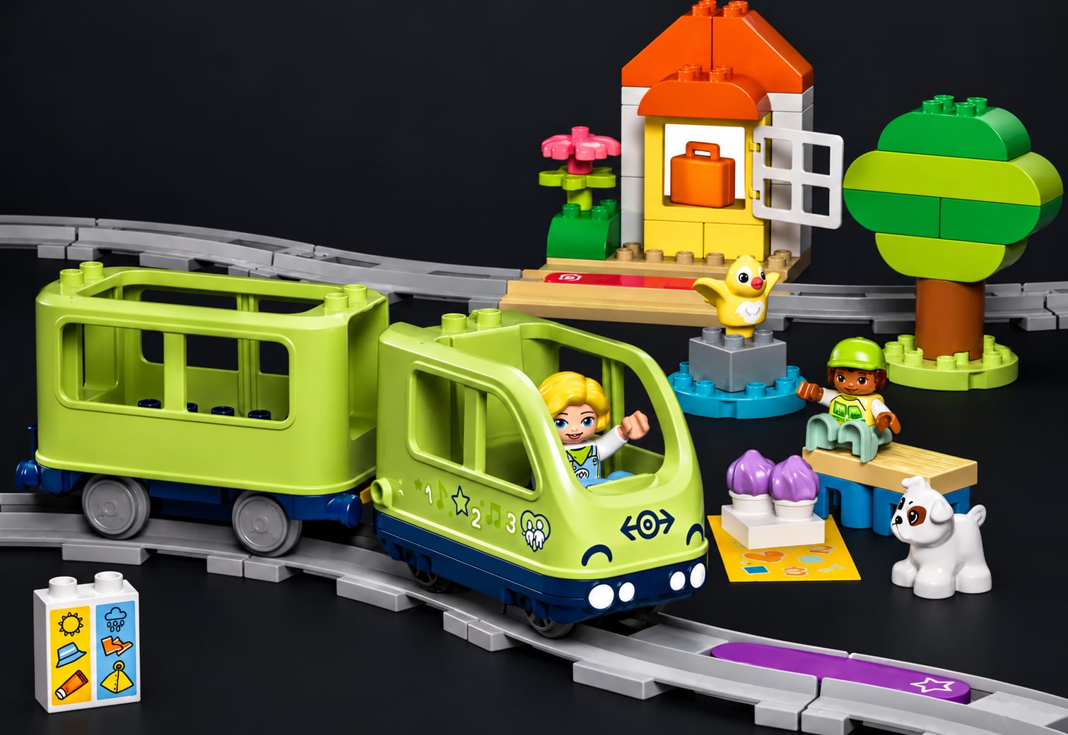 A green LEGO DUPLO train with a driver on curved track, next to a small station, a tree and figures